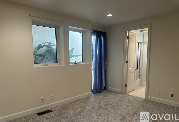 A room with a carpeted floor, two windows with blue curtains, and a door leading to another room.
