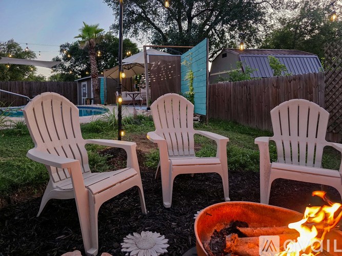 Three white chairs are placed in a backyard with a fire pit in the foreground.