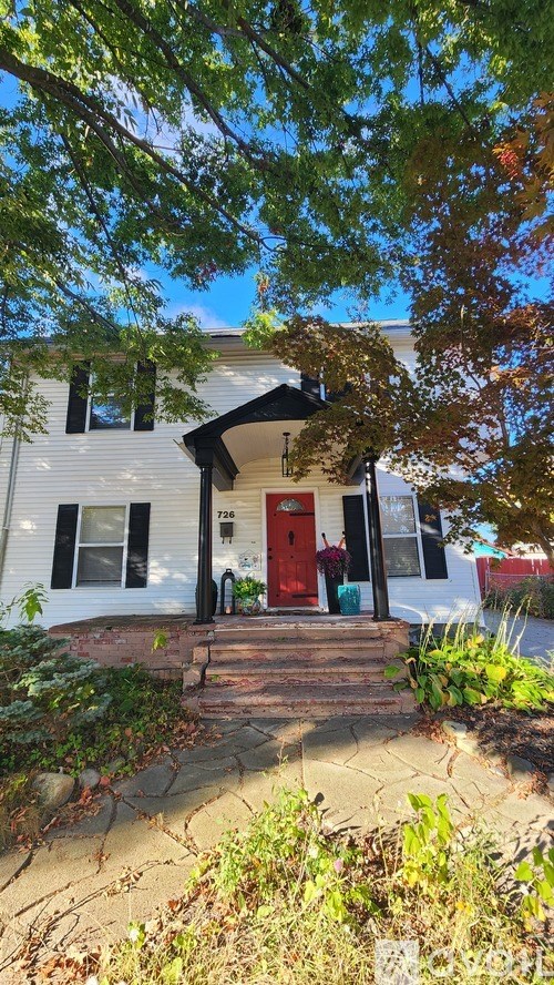 A white house with a red door is surrounded by greenery.
