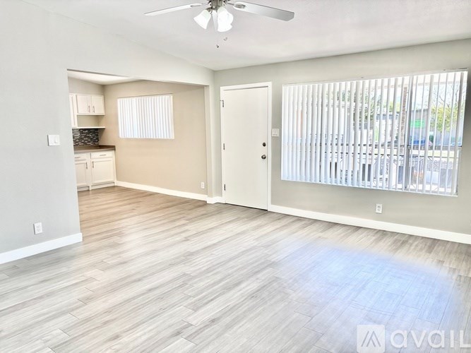 A spacious room with a ceiling fan and light, hardwood flooring, and a view of a parking lot through the window.