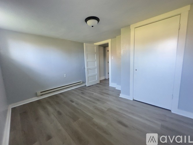 A room with a light wood floor and white walls, with a door on the right and a window on the left.
