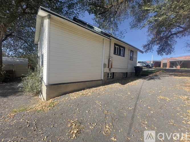 A small, single-story house with a garage door is situated in a driveway with trees and a clear sky in the background.