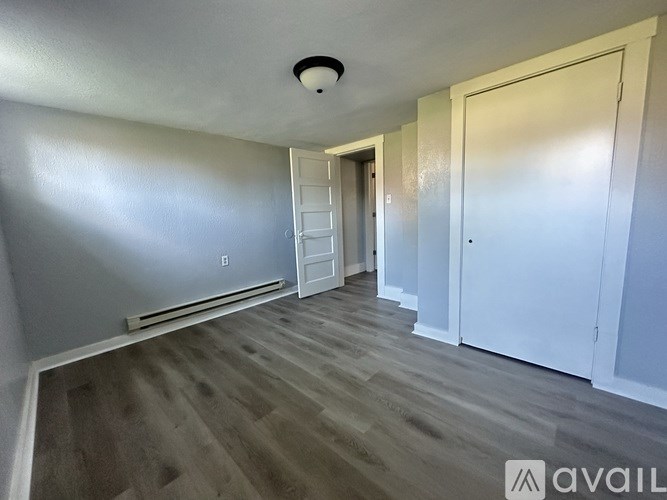 A room with a wooden floor and white walls, with a door on the right and a window on the left.