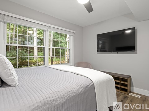 A bedroom with a bed, a window with blinds, a flat screen TV on the wall, and a wooden nightstand.