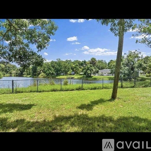 A grassy area with a fence and a tree in the foreground and a house and a body of water in the background.