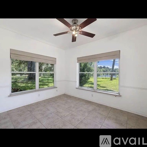 A room with a ceiling fan and two windows overlooking a green area.