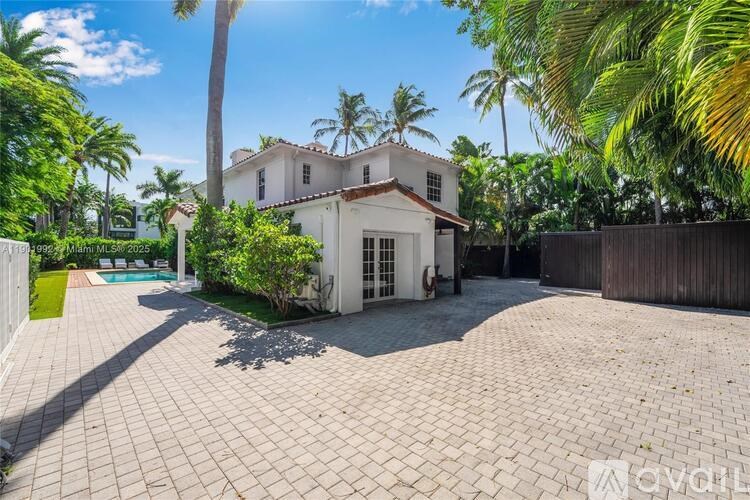 A house with a white exterior and a red roof is surrounded by a driveway and palm trees.