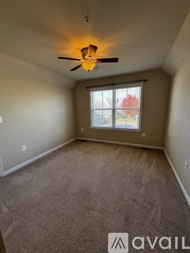 A room with a carpeted floor, a ceiling fan, and a window showing an outdoor view.