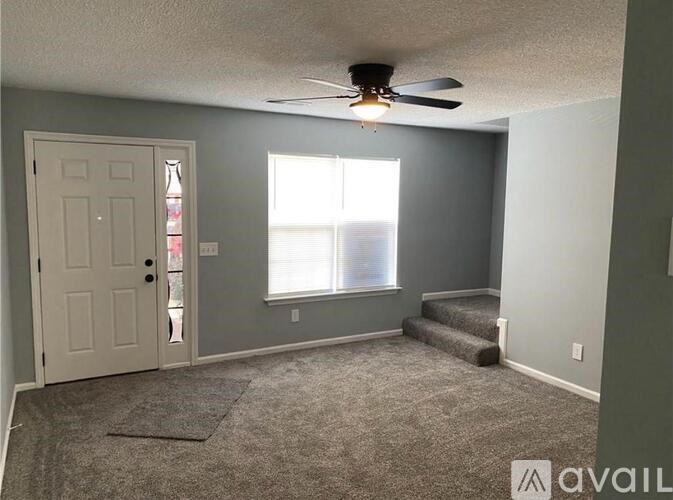 A room with a carpeted floor, a ceiling fan, and a door leading to another room.