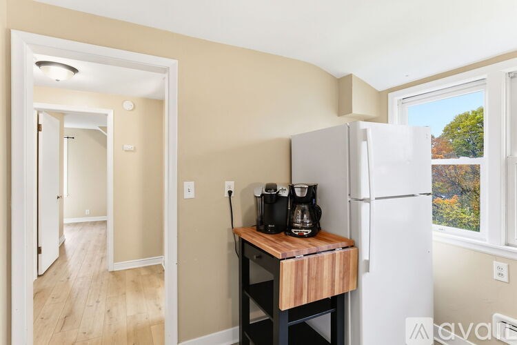 A kitchen area with a white fridge, a wooden table with a coffee maker on it, and a window showing trees outside.