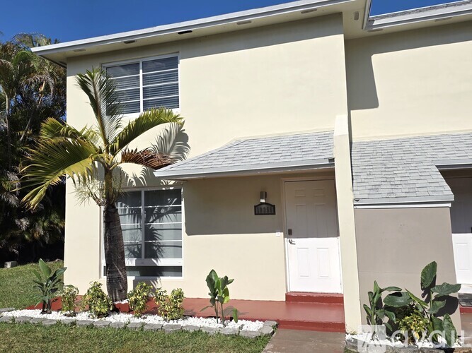 A house with a white exterior and a grey roof is surrounded by a green lawn and palm trees.