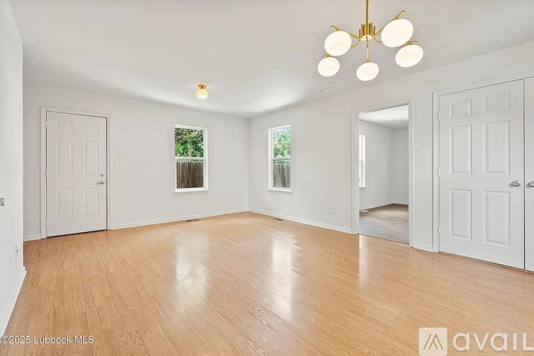 A spacious room with wooden floors and white walls, featuring a chandelier and a window with a view of a fence and greenery.