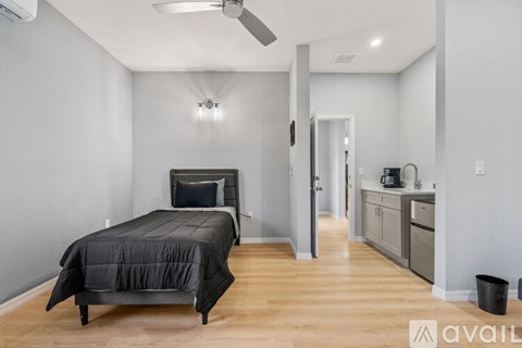 A bedroom with a bed, a ceiling fan, and a kitchen area in the background.