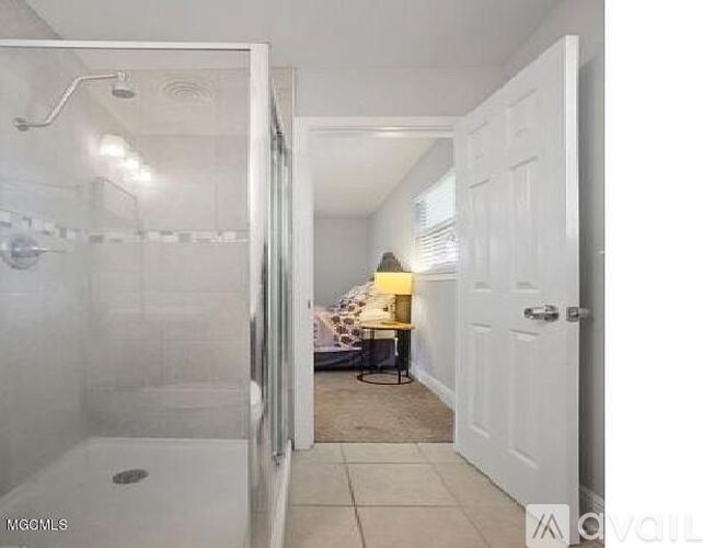 A white bathroom with a glass shower door and a white door leading to a bedroom.