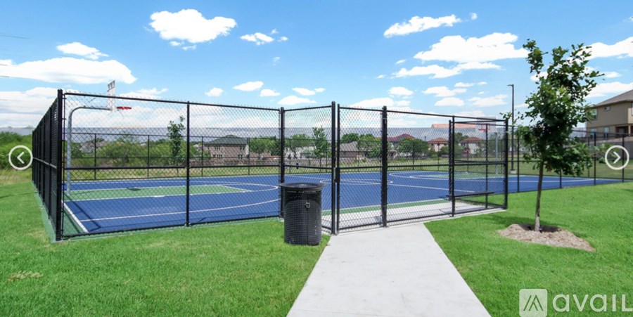 A tennis court is enclosed by a black fence and has a trash can and a walkway leading to it.