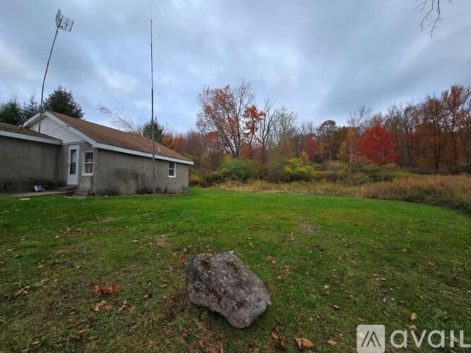 A large rock sits in the foreground of a grassy field with a house and antenna in the background.