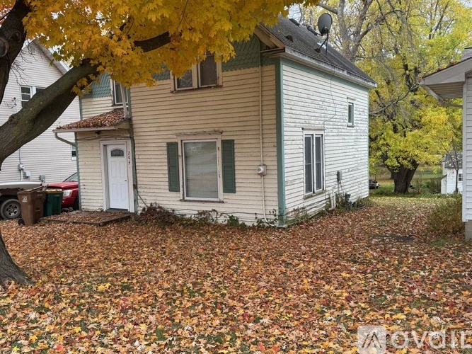 A house with a white door and green shutters is surrounded by fallen leaves.