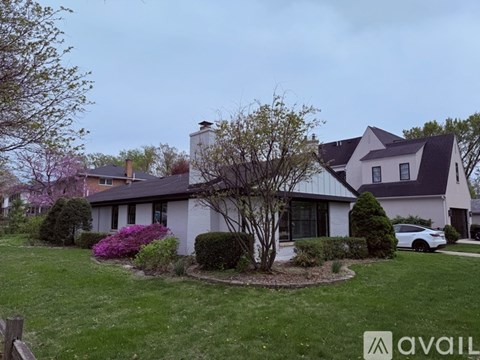 A house with a white exterior and a grey roof is surrounded by a green lawn and a small tree with purple flowers.