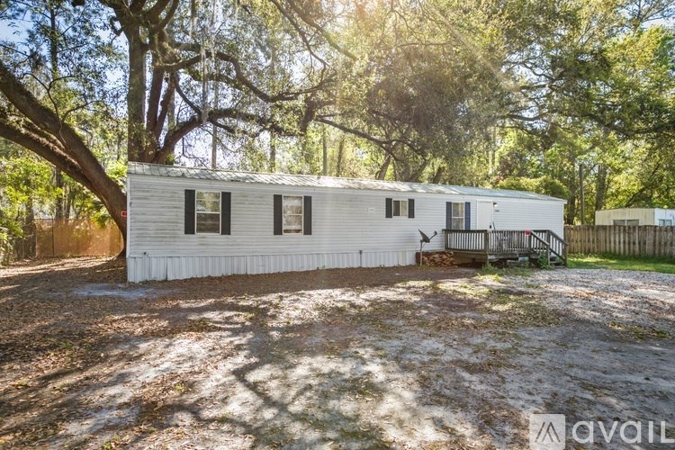 A white mobile home is surrounded by trees and has a gravel driveway.