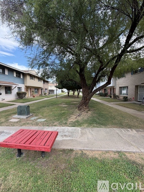 A red bench sits in the middle of a grassy area with a tree and apartment buildings in the background.