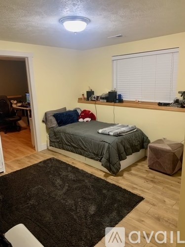 A bedroom with a bed, a window with blinds, a desk with a computer, and a rug on the floor.