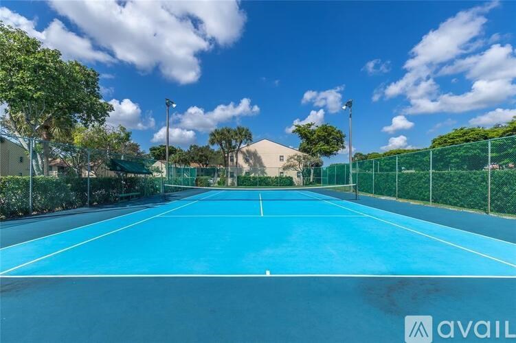 A tennis court with a blue surface and white lines, surrounded by a green fence and trees.