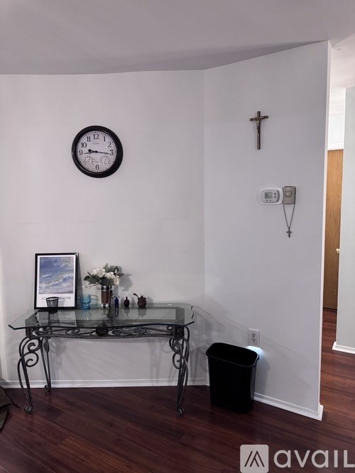 A black clock on a white wall with a cross and a picture frame on a glass table.