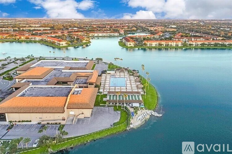 A large building with a swimming pool is surrounded by water and has a parking lot in front.