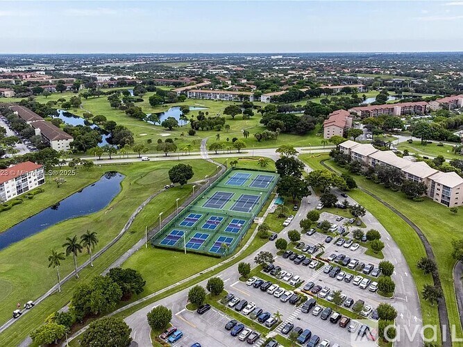 A large green field with a tennis court in the middle surrounded by a parking lot.