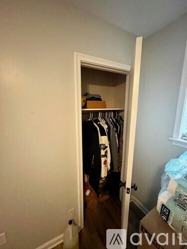 A small closet with clothes hanging up and a box of toilet paper on the floor.