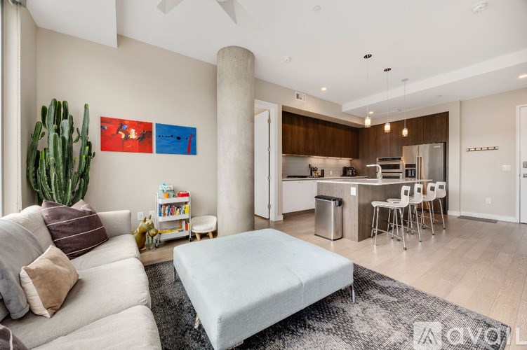 A living room with a couch, a chair, a bookshelf, and a kitchen area in the background.