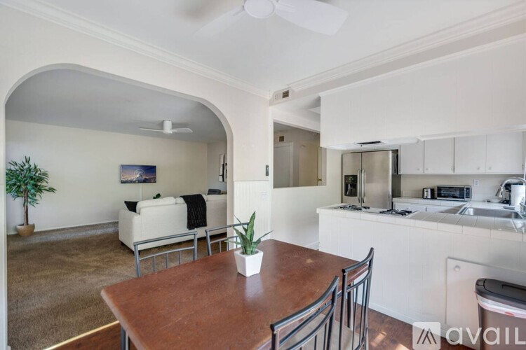 A kitchen with a table and chairs in the foreground and a living room with a couch and a plant in the background.