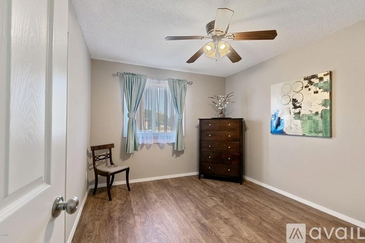 A room with a ceiling fan, a chair, a window with curtains, and a painting on the wall.