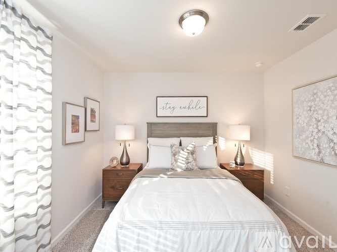 A bedroom with a bed, two nightstands, and a framed sign that says "stay a while.".