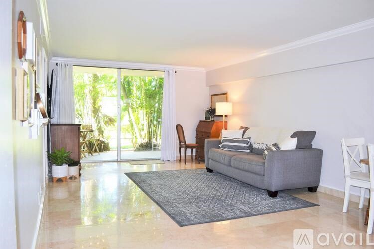 A living room with a grey couch, a chair, a table with a lamp, and a large window with a view of greenery outside.
