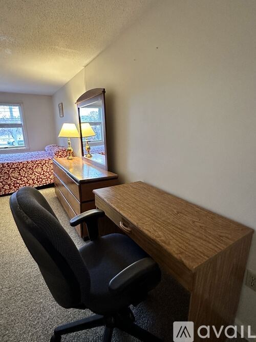 A black chair is in front of a wooden desk with a lamp on it.