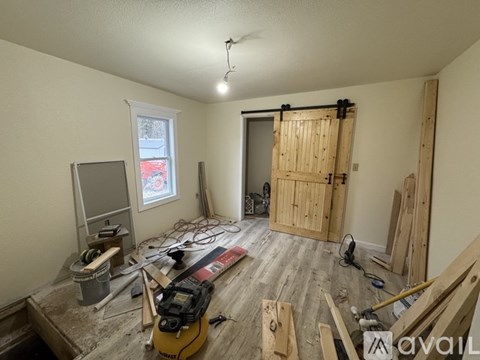 A room under construction with wood planks on the floor and a window with a view of a red car and trees outside.