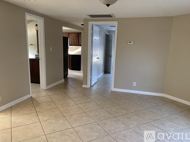 A spacious room with beige walls and tiled flooring, leading to another room with a door.
