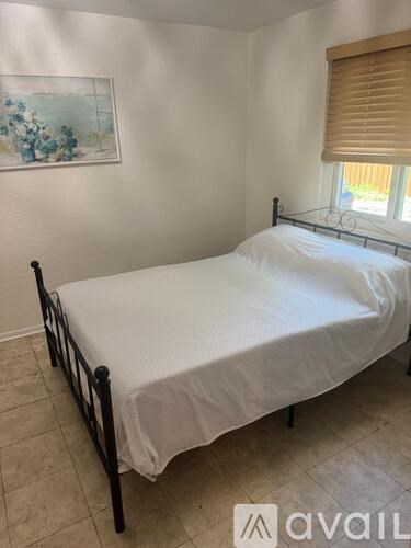 A bed with white bedding sits in a room with a painting on the wall and a window with blinds.