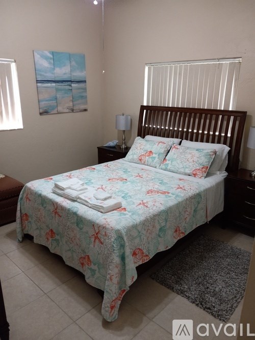 A bed with a floral patterned bedspread is in a room with a painting on the wall and a window with blinds.