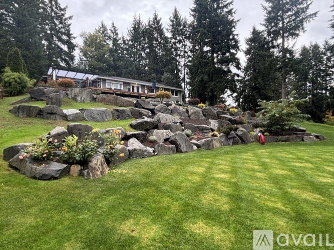 A large rock formation is in the foreground of a grassy area with a house and trees in the background.