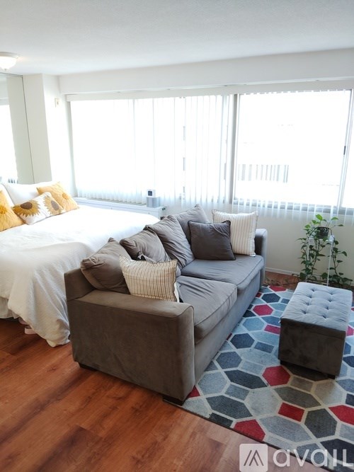 A grey couch with a white pillow is in the middle of a room with a bed in the background.