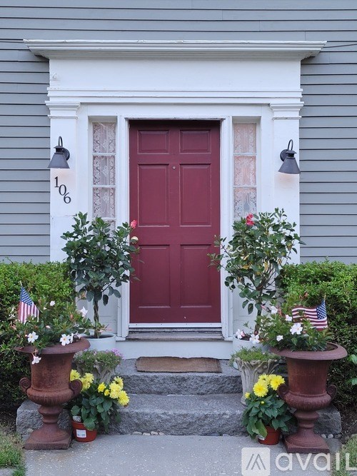 A red door is flanked by two planters with flowers and American flags.