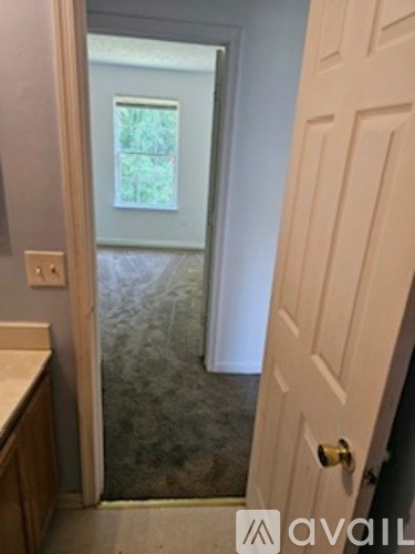 A white door with a gold handle is in the foreground of a bathroom with a window and a shower.