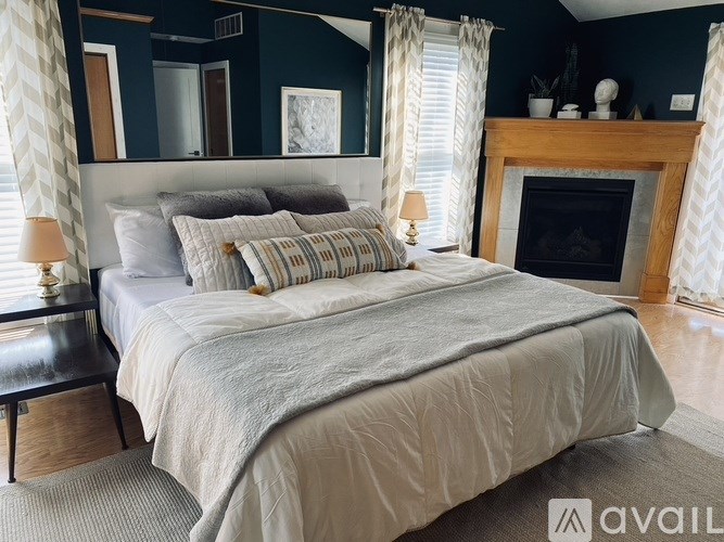 A bed with a grey comforter and pillows is in the foreground of a room with a fireplace.