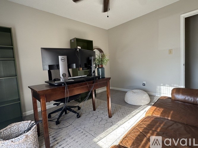 A room with a brown leather couch, a wooden desk with a computer and a plant on it, and a white ottoman.