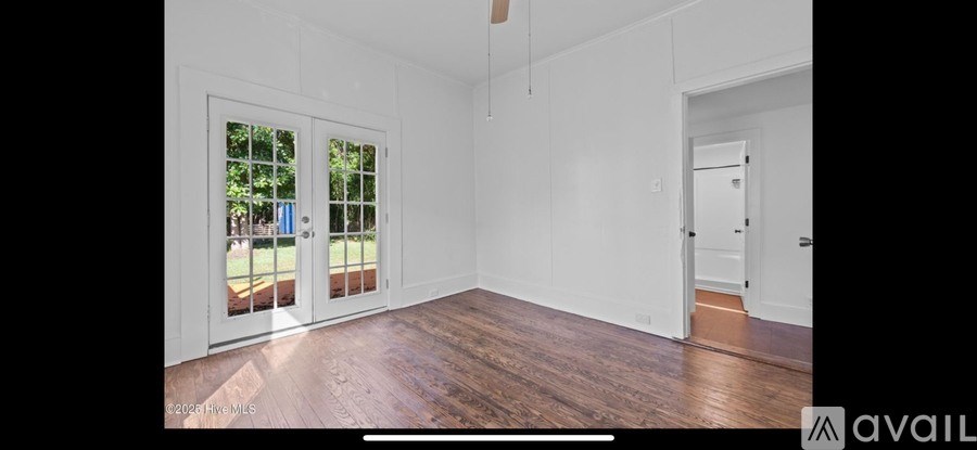 A spacious room with white walls and wooden flooring, featuring a large window with a view of a green lawn.