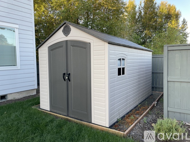 A storage shed with a grey door and windows is situated in a grassy area.