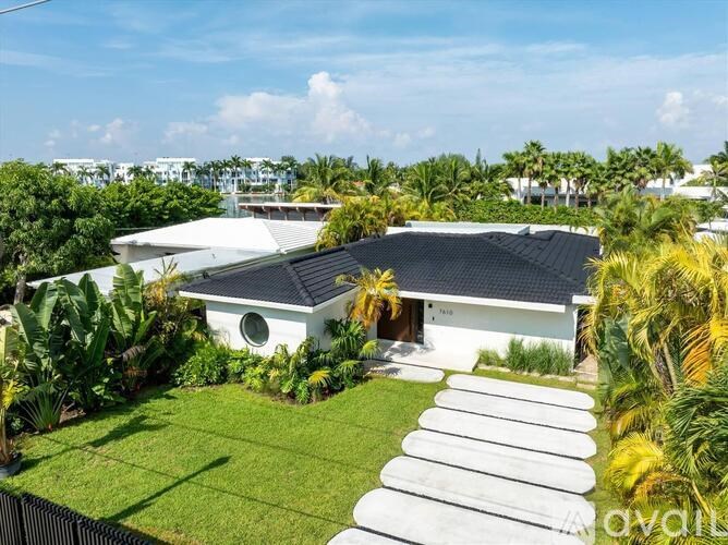 A white house with a black roof surrounded by greenery and a clear blue sky.