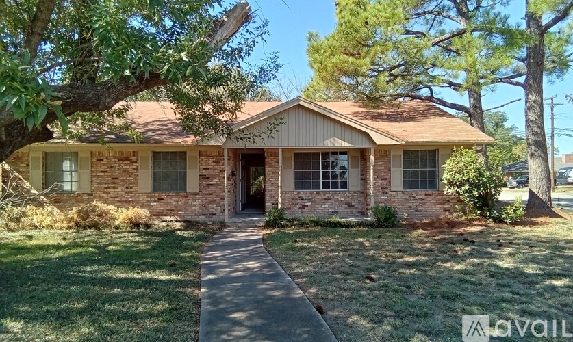 A house with a brown roof and a brick wall is surrounded by green grass and trees.